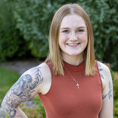 Portrait of Kendall Mara. She is standing in a garden and smiling at the camera. She has a black and white tattoo sleeve. 