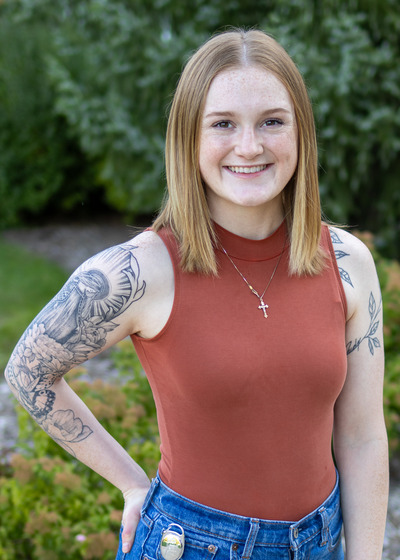 Kendall Mara smiles at the camera. She is standing in a garden and is wearing a tank top and has a black and white tattoo sleeve.