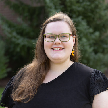 Morgen Clark smiles at the camera. She has long brown hair, green glasses, and orange earrings. She is standing outdoors in front of blurred evergreen trees. 