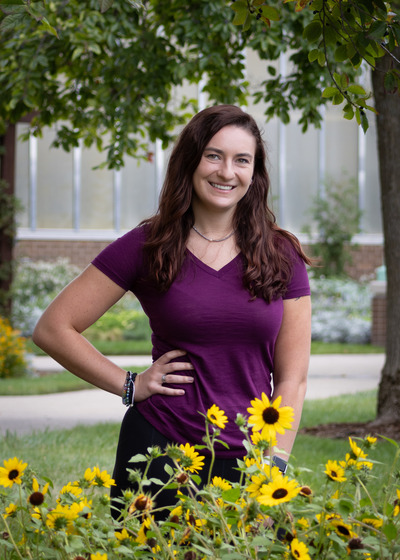 Rosemary Northcote stands with one hand on her hip behind some sunflowers in the MSU gardens.
