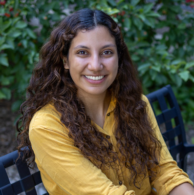 This image shows Sarah Gonzalez Henao sitting on a bench and smiling at the camera. She is wearing a mustard yellow shirt and has long curly hair. 