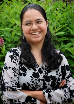 A person with long brown hair has their arms crossed and is smiling at the camera. They are standing in front of a shrub. 