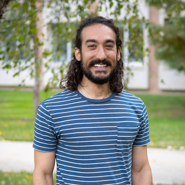 Dr. Alexander Mela stands outside and smiles at the camera. He's wearing a blue and white striped shirt and jeans and has long curly hair and a beard and mustache. He looks friendly.