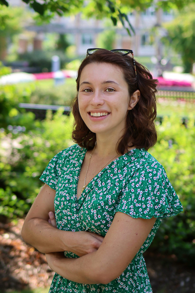 Camille Boucaud stands in a garden smiling at the camera. She's wearing a green and white dress and is crossing her arms.
