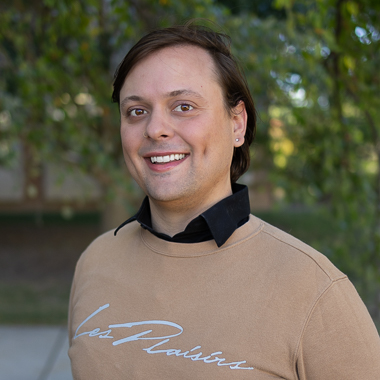 Joel Marty smiles at the camera. He's wearing a beige sweatshirt that reads "Les Plaisirs" over a black collared shirt. 