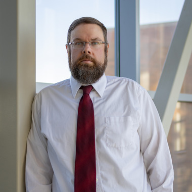 Phil Delekta looks at the camera. He is wearing a white shirt and a red tie. He has a bear, glasses, and short brown hair. He is standing in front of windows. 