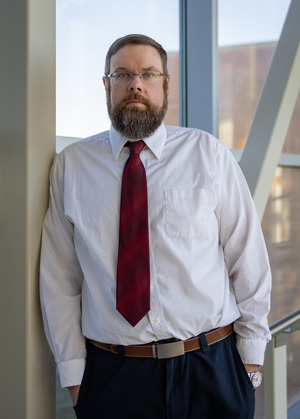 Dr. Phillip Delekta looks at the camera. He is wearing a white shirt and red tie and has a beard and short brown hair. He is standing in front of windows. 