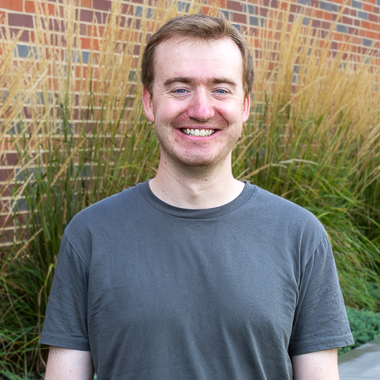 Jared Godfrey stands outside in front of grasses and is smiling at the camera. He's wearing a dark gray t-shirt and has short hair. 