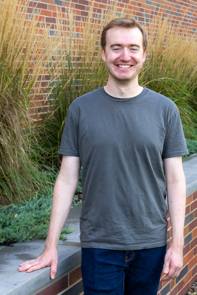 Jared Godfrey stands in front of grasses and next to a low brick wall. He's smiling at the camera. He's wearing a dark gray shirt and jeans and has short hair.