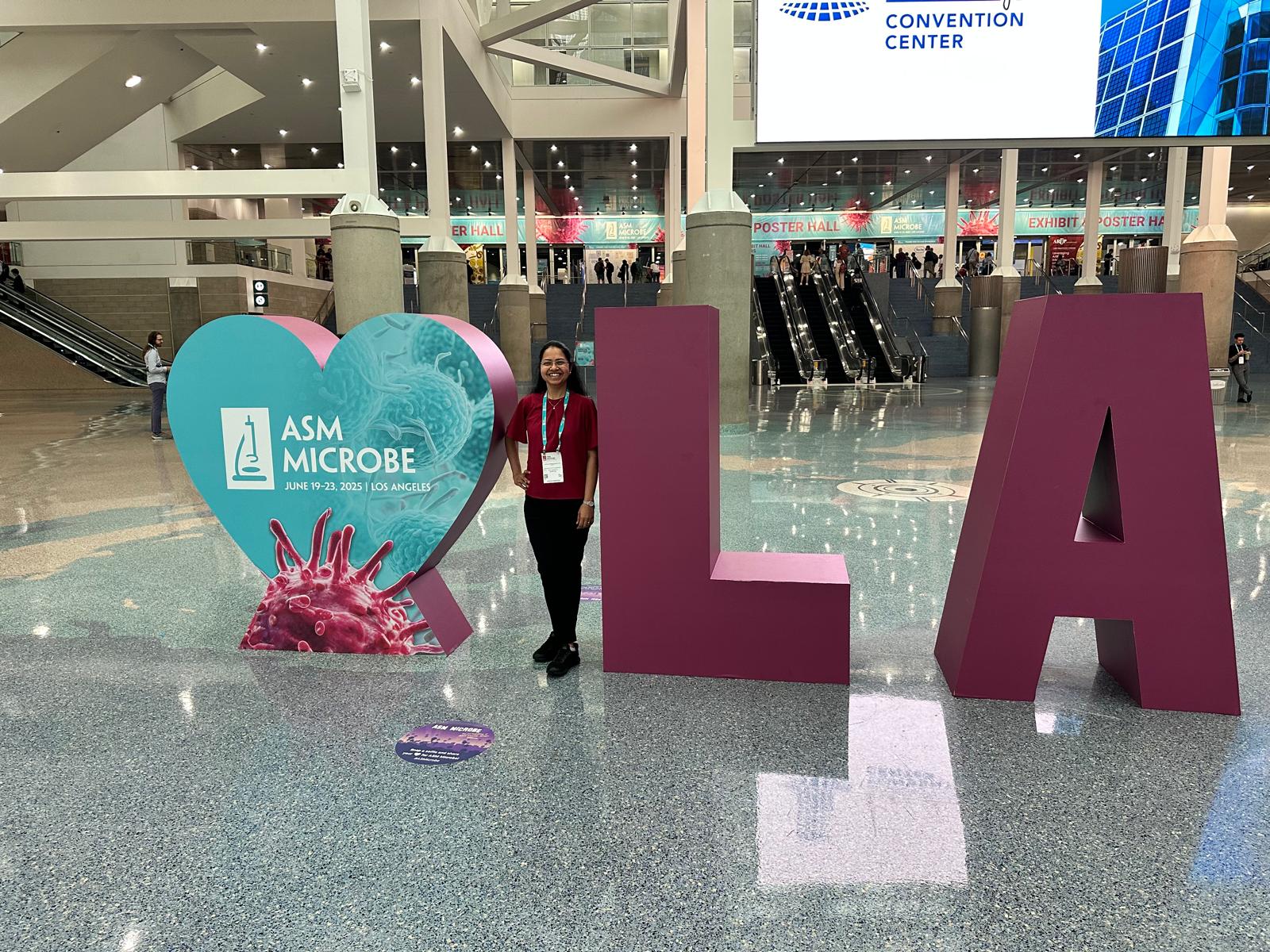 Dr. Rangarajan stands next to a heart-shaped sign reading "ASM Microbe." On her other side are a large L and A.