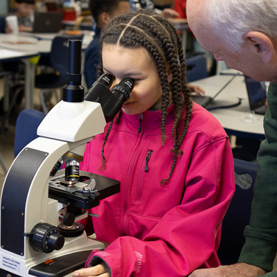 An elementary student looks through a microscope while Dr. Hardy looks on. 