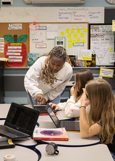 Djeneba Diallo points to a computer while two girls look on. They are in an elementary school classroom.