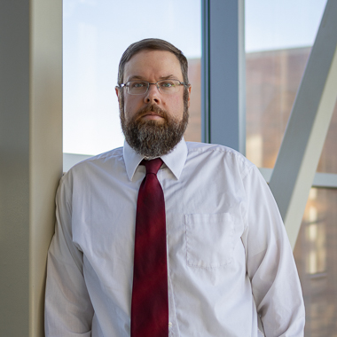 Phil Delekta looks at the camera. He is wearing a white shirt and a red tie. He has a bear, glasses, and short brown hair. He is standing in front of windows. 