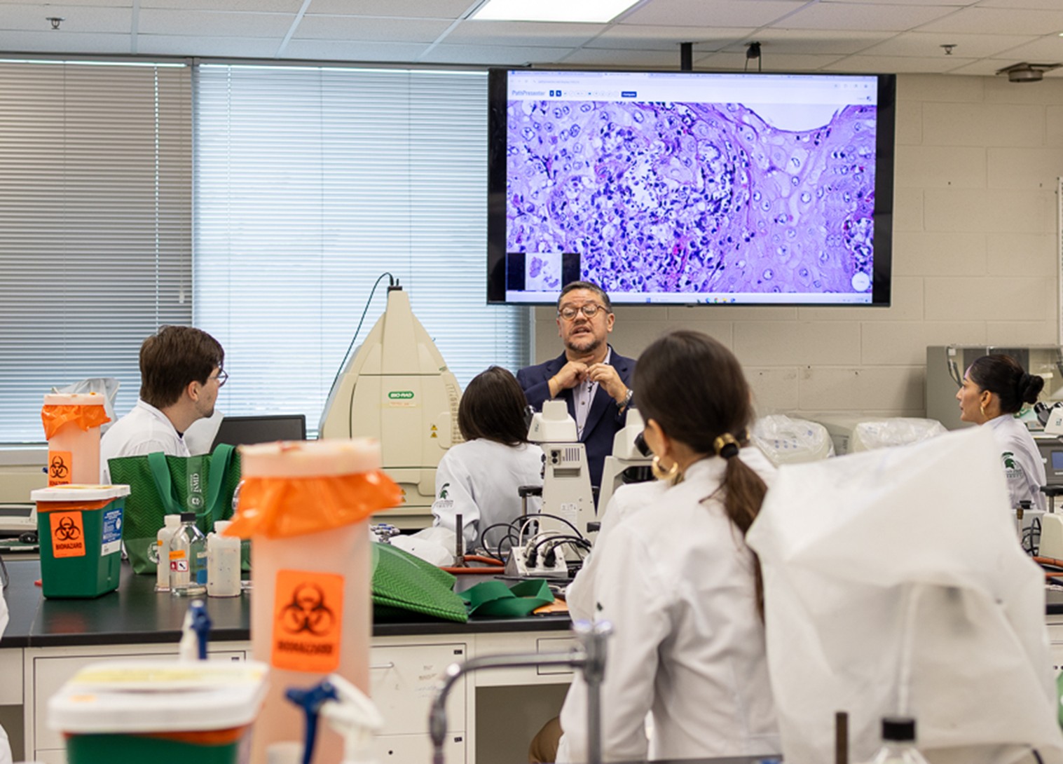 Dr. Laga stands at the front of the teaching lab and gestures while teaching. He is standing in front of a TV that is showing a purple-stained cell.