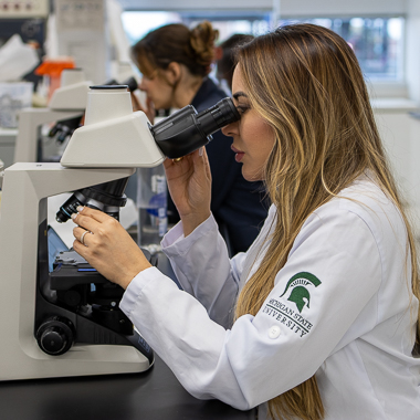 Woman in MSU lab coat looks through microscope.