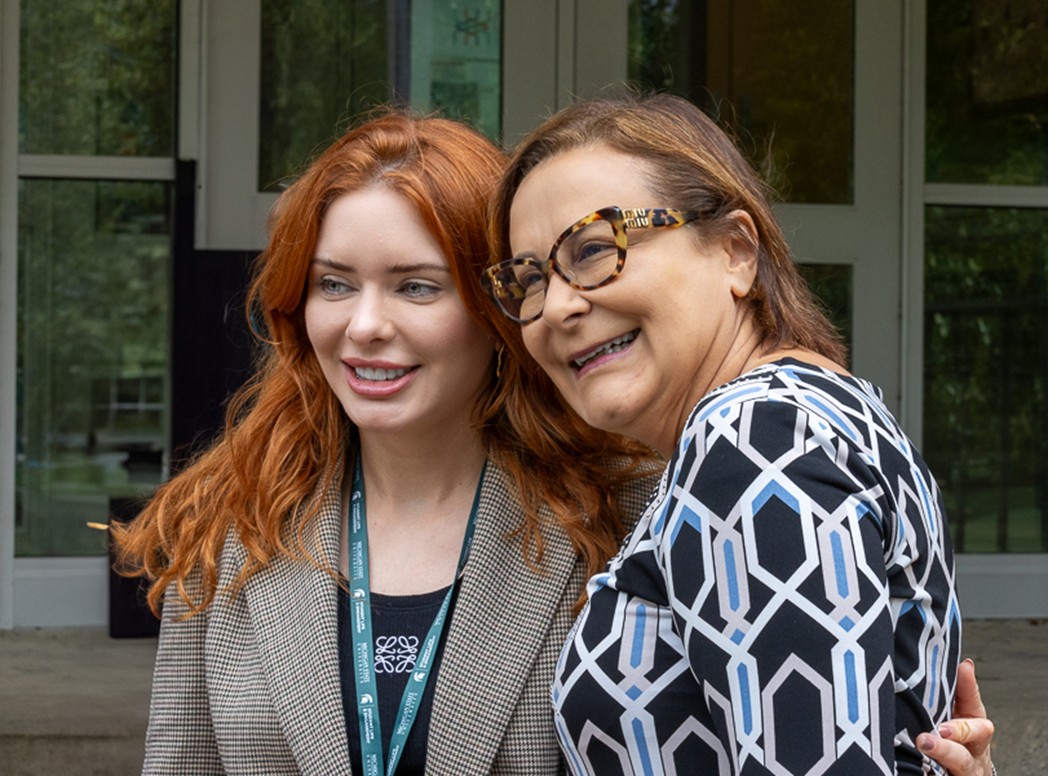 Two woman stand together and smile at a camera.