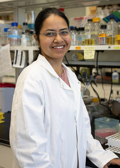 Aathmaja Anandhi Rangarajan stands at her lab bench. She is wearing a lab coat and glasses and is smiling at the camera.
