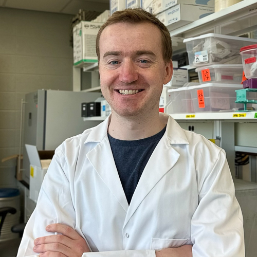 Jared Godfrey smiles at the camera. He is wearing a lab coat and standing in his lab. 