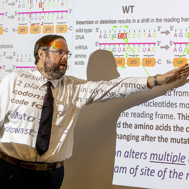 Dr. Phillip Delekta, wearing a white shirt, dark tie, and glasses gestures toward a projected slide while teaching a genetics class. The slide displays diagrams and explanations of genetic mutations and reading frames. The projection is also seen on him - his shirt and face.