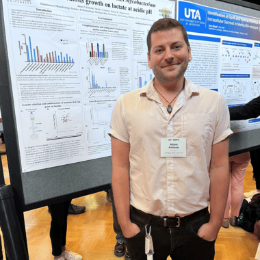 Adam Kibiloski stands in front of his poster at the Midwest Microbial Pathogenesis Conference. He's wearing a short-sleeve buttondown shirt and dark pants and has a name tag on. 