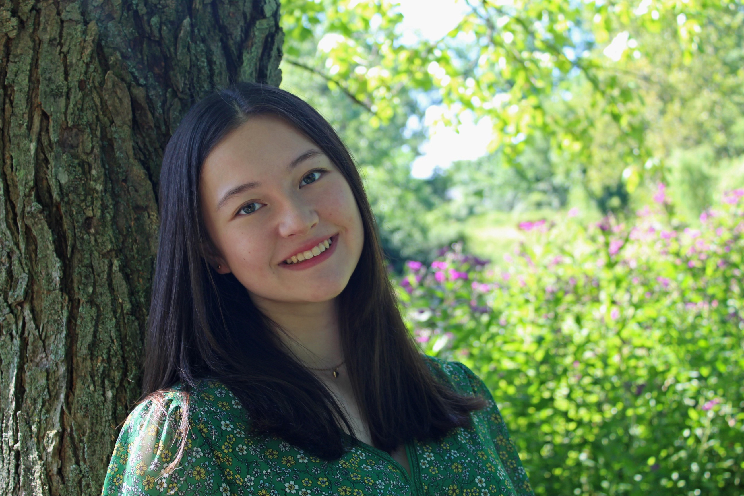 Girl with long black hair smiles at the camera. She is leaning against a tree. There are pink flowers in the background. 