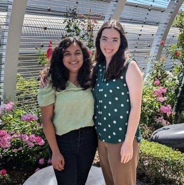 Ashwini Ramesh and Abigail Lippert stand next to each other and smile at the camera. They are standing in front of flowers and rock-shaped benches on a terrace. 