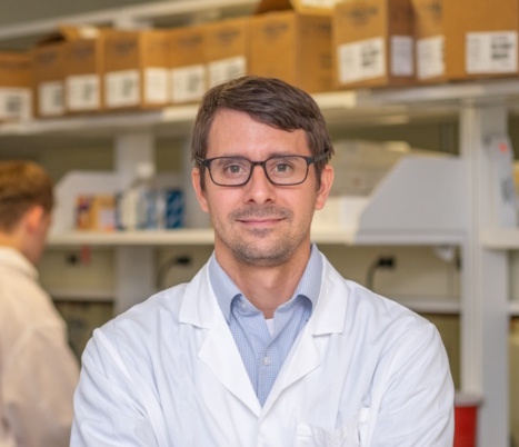 Man wearing glasses looks straight into the camera. He is wearing a lab coat and standing in front of a lab bench.