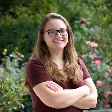 The image shows Beth Ottosen with long, light brown hair wearing glasses and a maroon short-sleeved shirt, standing outdoors. She is smiling and has her arms crossed. The background features a lush garden with various green shrubs and blooming flowers, including pink and red roses.