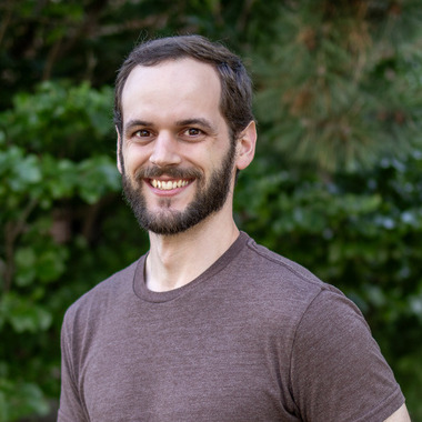 Troy Burtchett smiles at the camera. He is wearing a brown shirt and has a beard and moustache. He is standing in front of evergreen trees. 