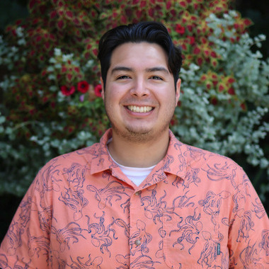 Jasper Gomez smiles at the camera. He is wearing a coral colored shirt with blue octupuses all over it, and he is standing in front of colorful plants. 