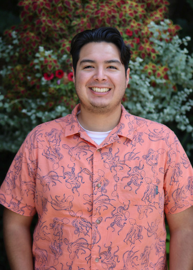 Jasper Gomez smiles at the camera. He is wearing a coral colored shirt with blue octopuses all over it. He is standing in front of brightly colored plants. 