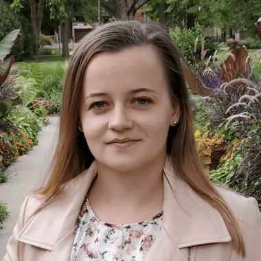 Woman in floral shirt and pink jacket smiles serenely at the camera. She is standing in a garden. 