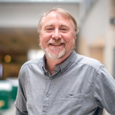 Rich Lenski smiles at the camera. He is standing in a hallway, but the background is blurred and not of consequence. He is wearing a chambray button down shirt. 