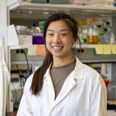 Lexi Vu is standing in a laboratory setting, smiling at the camera. She is wearing a white lab coat over a gray shirt. Behind her, a shelf is lined with various laboratory supplies, including bottles and containers in different colors, suggesting a well-equipped lab environment. There are sticky notes attached to the shelves, adding a touch of organization and personalization. The overall atmosphere is bright and professional.