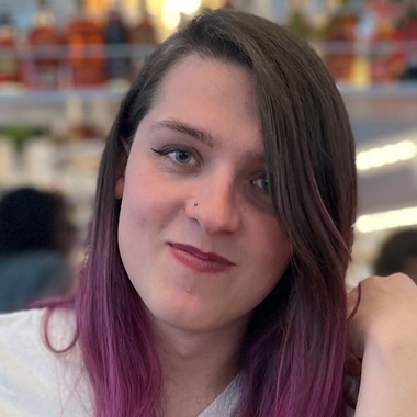 A person with long brown and pink hair smiles into the camera. She has sunglasses hanging from the front of her shirt.