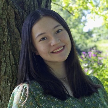 Girl with long black hair smiles at the camera. She is leaning against a tree. 