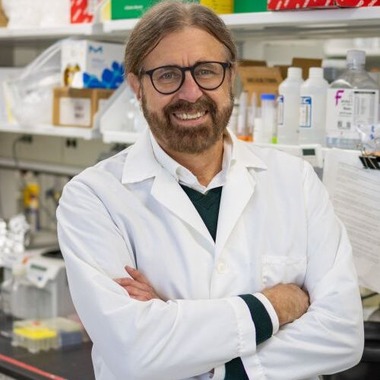 James Pestka smiles at the camera. He is wearing a lab coat and standing in front of a lab bench.