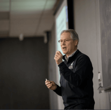 This image shows Dr. Brian Schutte standing and speaking in a classroom or lecture hall. He is wearing glasses and a dark sweater with a logo on the left side. He is holding a pen or a pointer in his right hand and appears to be engaged in explaining something. The background features a projector screen and a chalkboard or whiteboard. The room has a modern ceiling with recessed lighting.