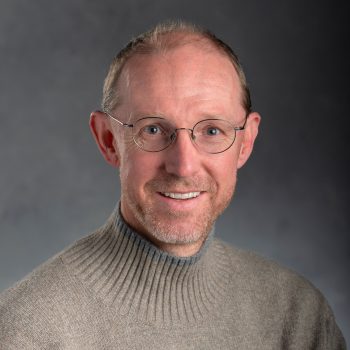 Sean Crosson smiles at the camera. He is sitting in front of a gray background wearing wire-rimmed glasses and a light brown sweater. 