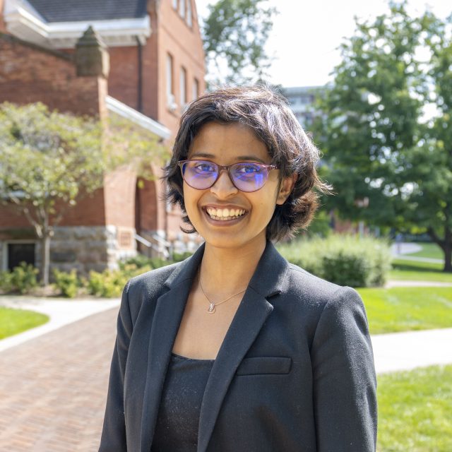 Veona Cutinho smiles at the camera. She has glasses on and a black suit jacket. She is standing in front of a beautiful brick building on MSU's campus. 