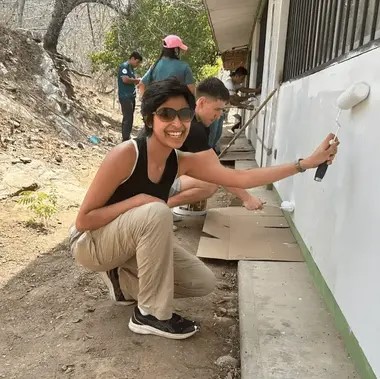 Veona Cutinho grins while painting a wall with a roller brush. She is kneeling and there are people behind her who are also painting. 