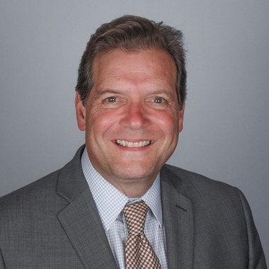 Victor DiRita smiles at the camera. He is wearing a gray suit and sitting in front of a gray background. 