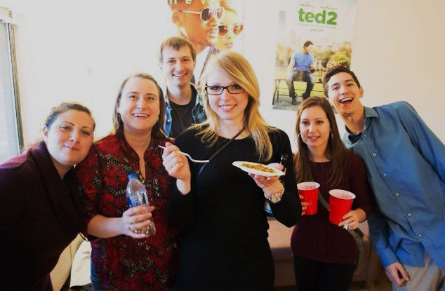 A group of people are smiling broadly at the camera - clearly having a great time. The girl in the center has a plate with food on it and a fork.