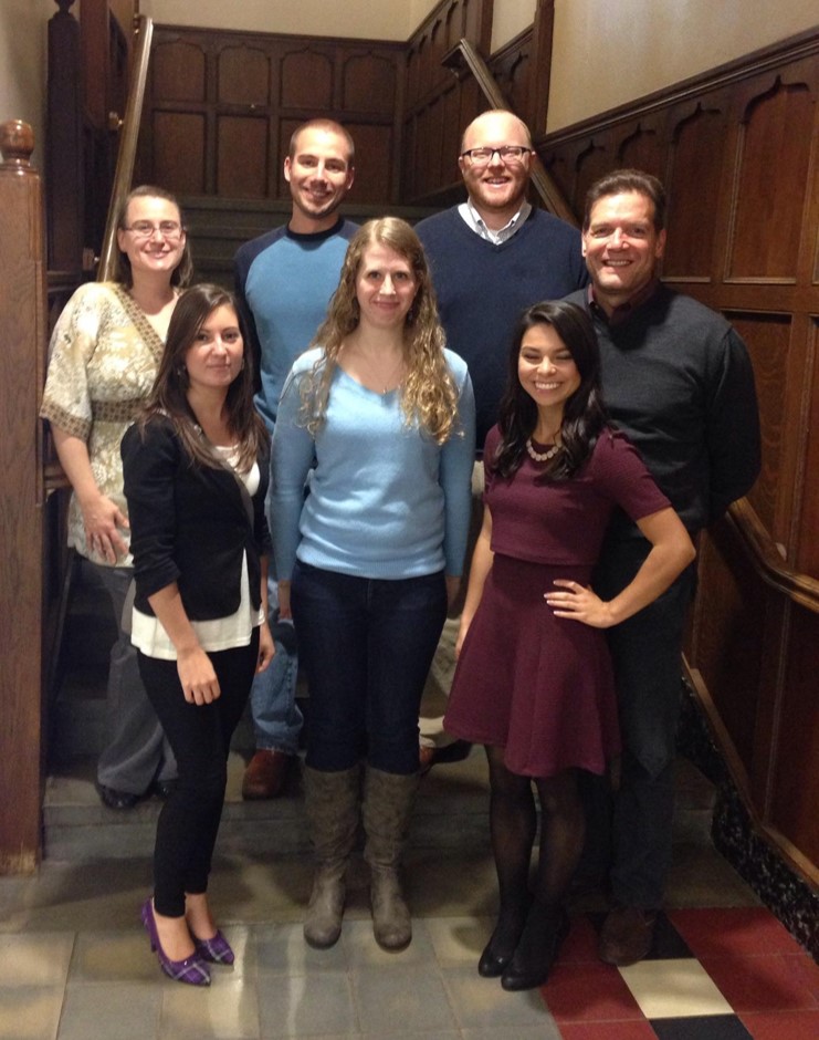 A group of people standing in a hallway. There is dark woodwork behind them.