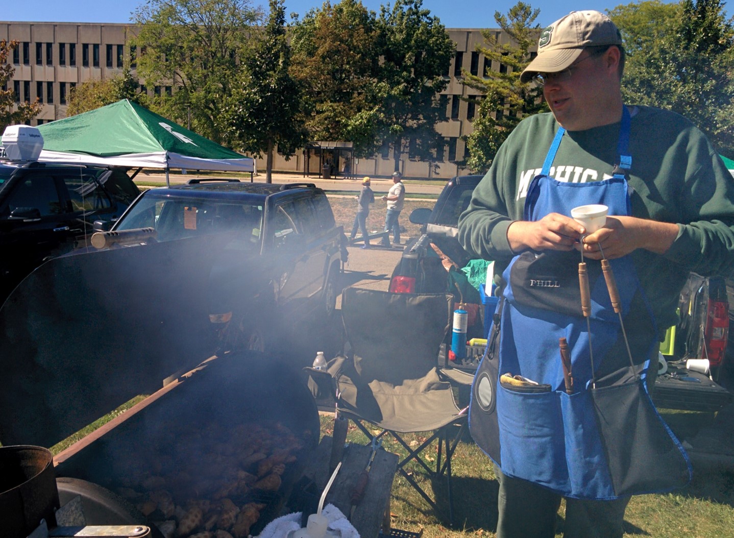 Phil stands next to the grill. He's wearing an apron and a baseball cap.