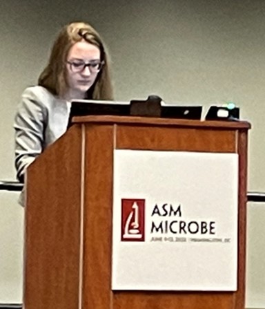 Beth standing behind a lecturn that reads ASM microbe.
