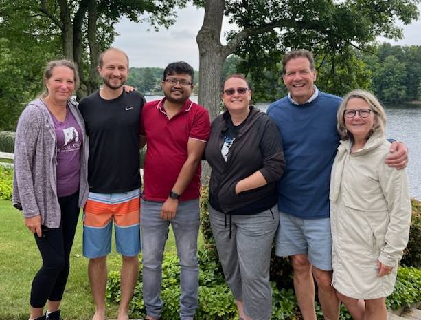 A group of people stand together. There is a lake and grass behind them.