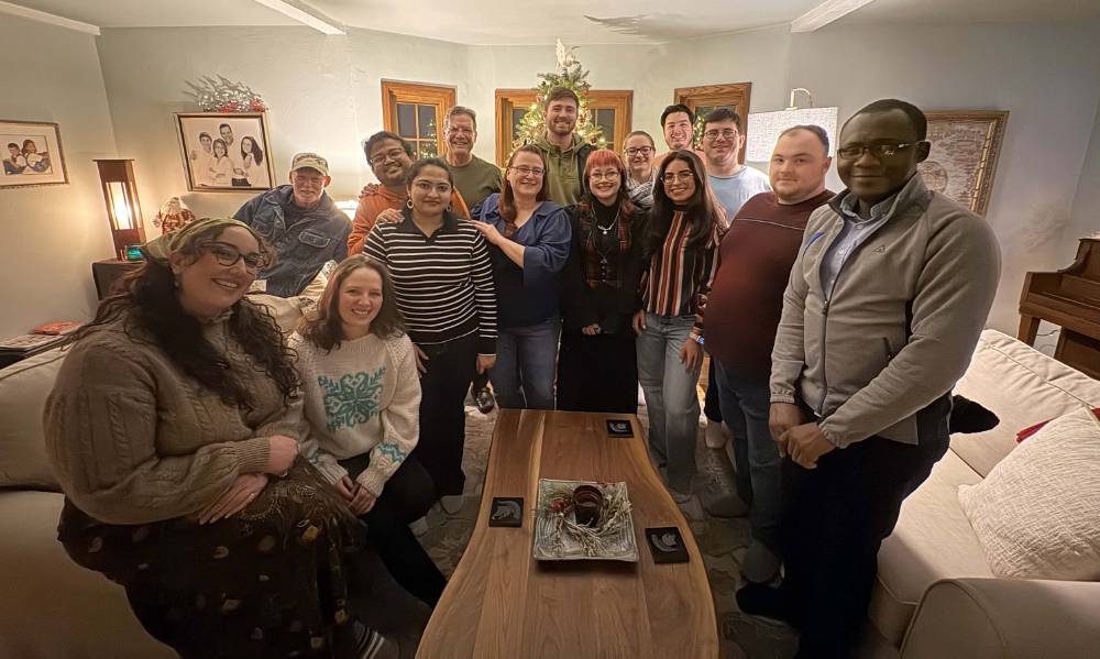 A group of people stand together in a party setting. The glow of a christmas tree is visible behind them.