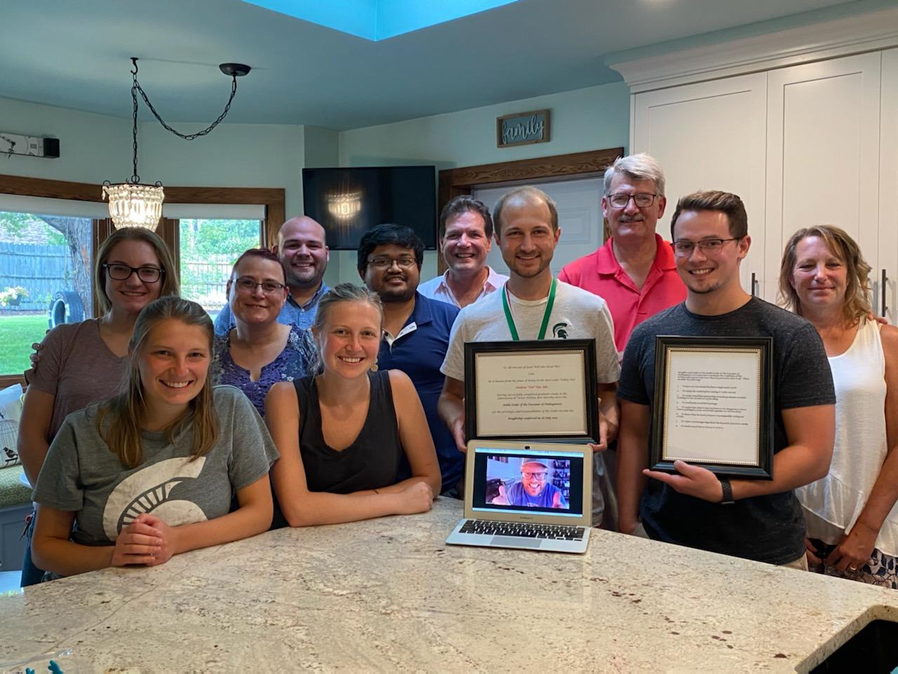 Group of people gathered around a kitchen island with a laptop. Two people are holding framed pictures.