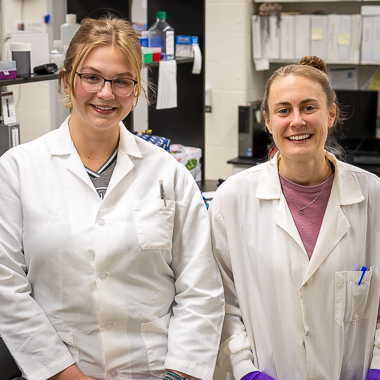 Two scientists stand next to each other. They are in lab coats and are standing in their lab. 